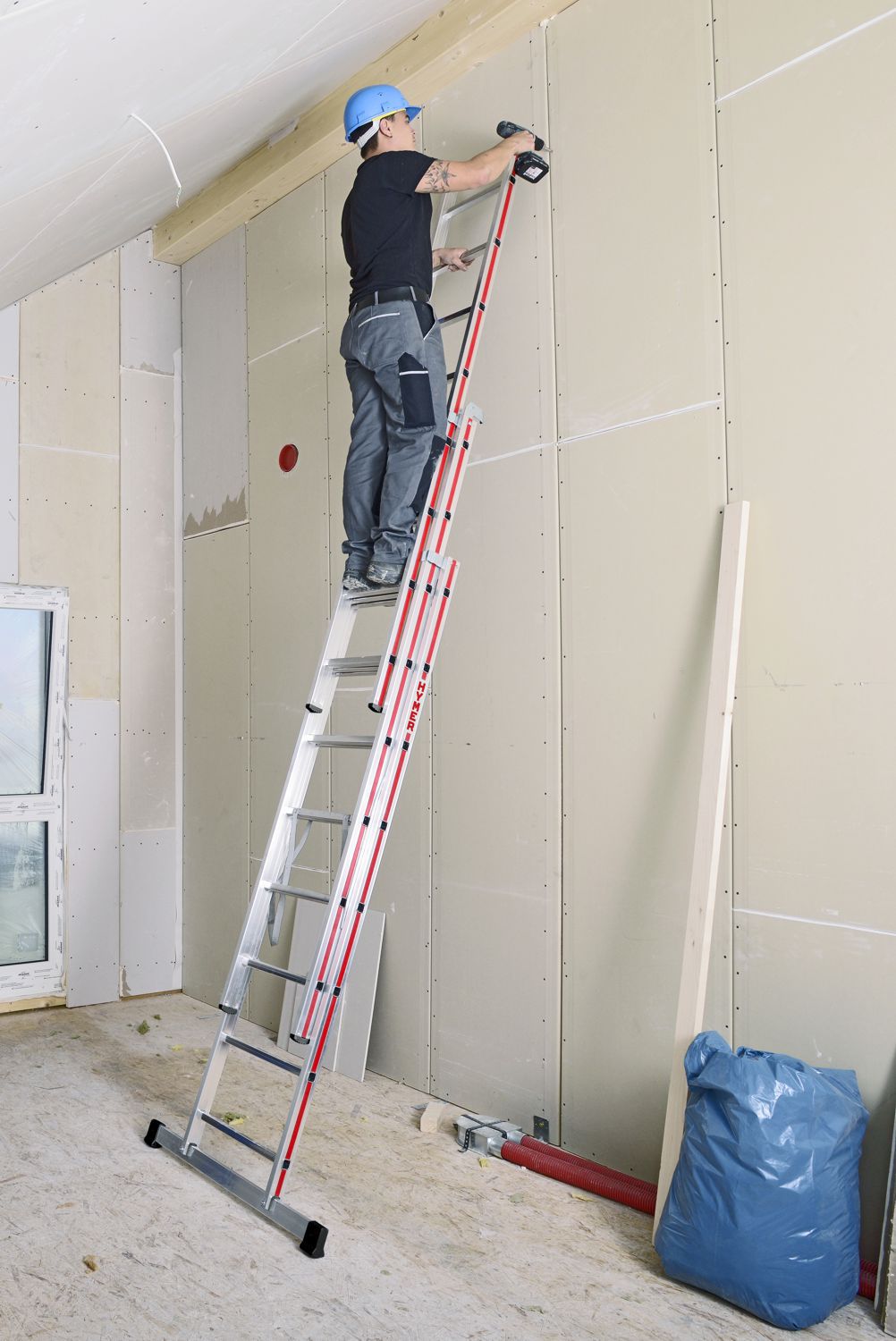 Handwerker auf einer dreiteiligen Mehrzweckleiter von Hymer stehend bei Arbeiten an einer Wand in einem Rohbau bild 2