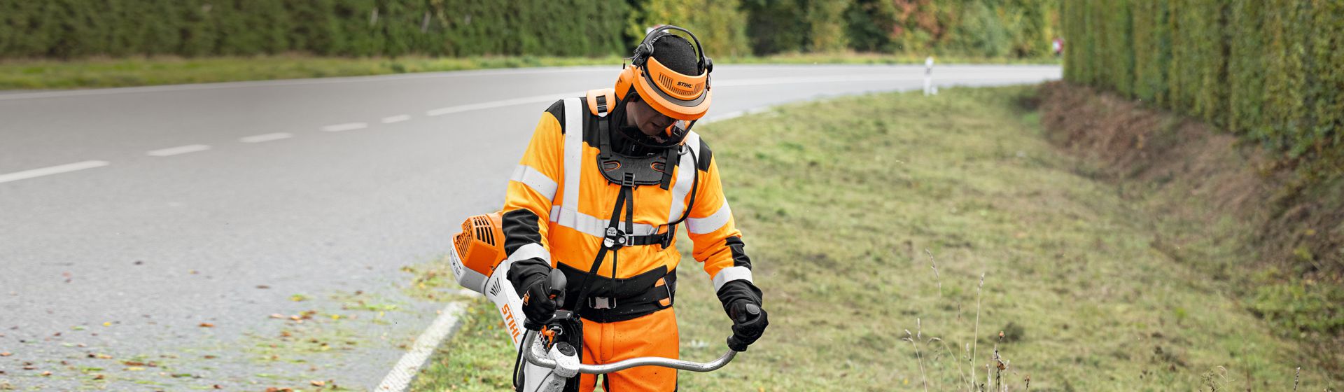 Mann in orangefarbener Stihl Warnschutzkleidung mit Schutzhelm bei Mäharbeiten an einer Landstraße bild