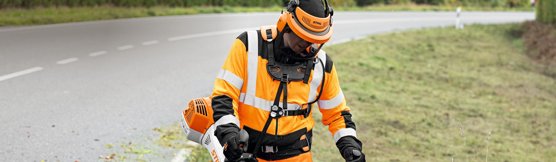 Mann in orangefarbener Stihl Warnschutzkleidung mit Schutzhelm bei Mäharbeiten an einer Landstraße bild