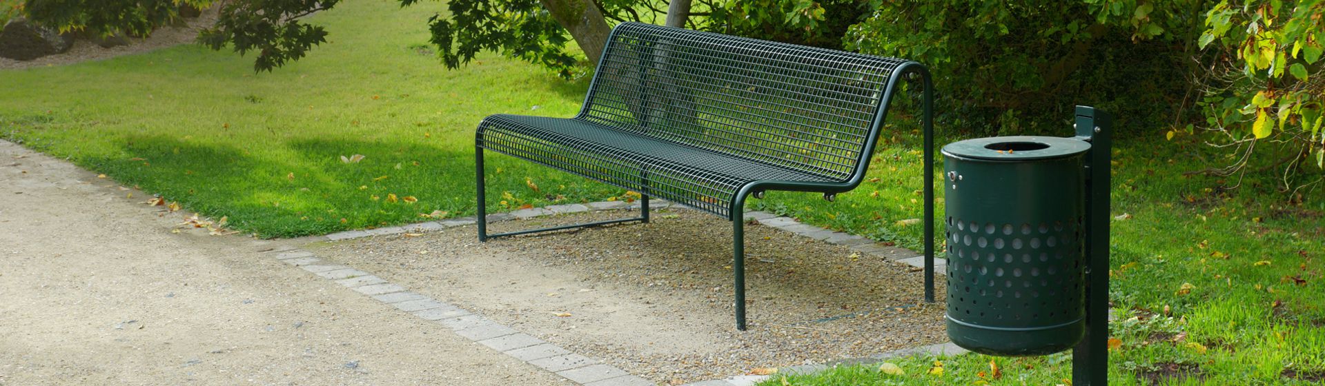 Dunkelgraue Bank mit Gitter Sitzfläche und Lehne auf einer Kiesfläche neben einem Kiesweg stehend, rechts daneben ein  dunkelgrauer Metall-Mülleimer, im Hintergrund Rasen und Sträucher bild