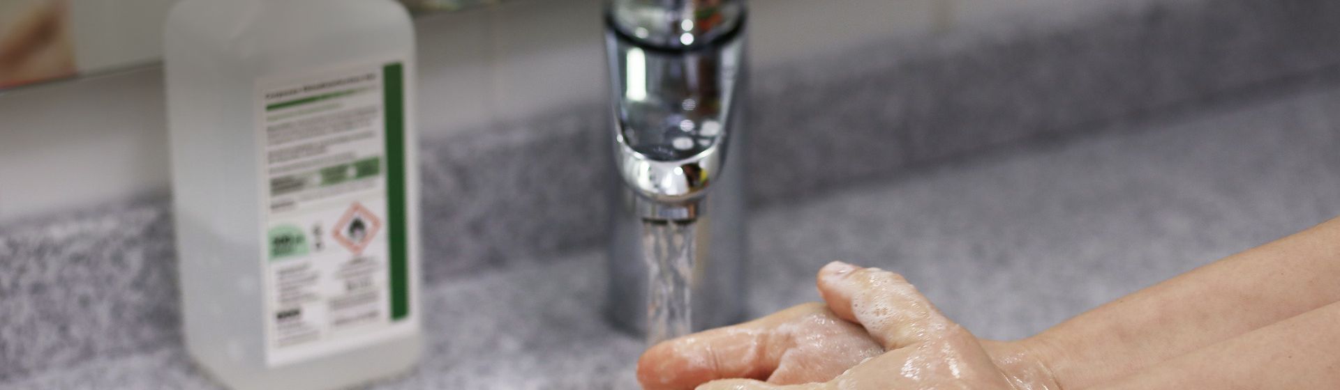 Händewaschende Person an einem Waschbecken, im Hintergrund ein Desinfetionsmittelspender auf dem Rand stehend bild