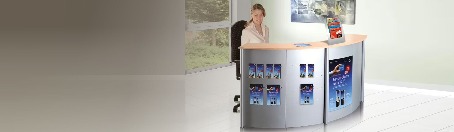 Eine Frau an einem halbrunden Empfangs-Thekensystem mit silberfarbenen Metallelemente und Holztheke sitzend, auf dessen Front Werbeplakate und Broschürenhalterungen angebracht sind bild
