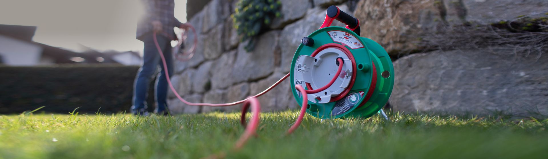 Grüne Brennenstuhl Garten-Kabeltrommel mit ausgerolltem roten Kabel stehend auf einer Rasenfläche vor einer Mauer bild