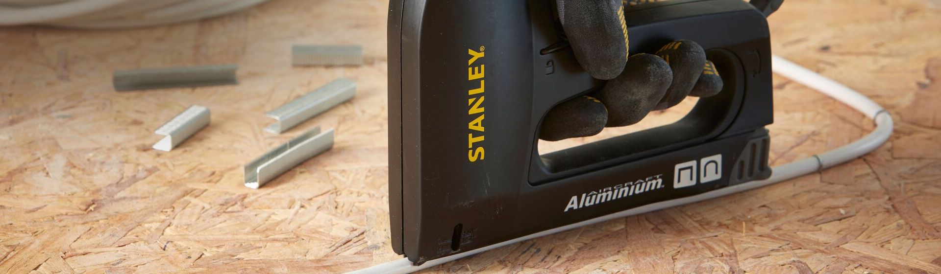 Schwarzer Handtacker der Marke Stanley beim Befestigen einer weißen Leitung auf einer Pressspanplatte bild